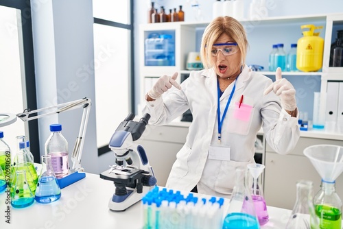 Middle age blonde woman working at scientist laboratory pointing down with fingers showing advertisement  surprised face and open mouth