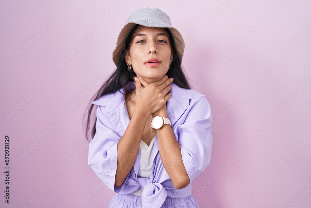 Young hispanic woman standing over pink background wearing hat shouting suffocate because painful strangle. health problem. asphyxiate and suicide concept. Young hispanic woman standing over pink background wearing hat shouting suffocate because painful strangle. health problem. asphyxiate and suicide concept.