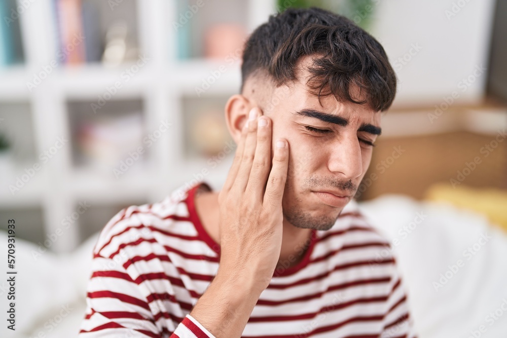 Young hispanic man suffering for ear pain sitting on bed at bedroom Young hispanic man suffering for ear pain sitting on bed at bedroom
