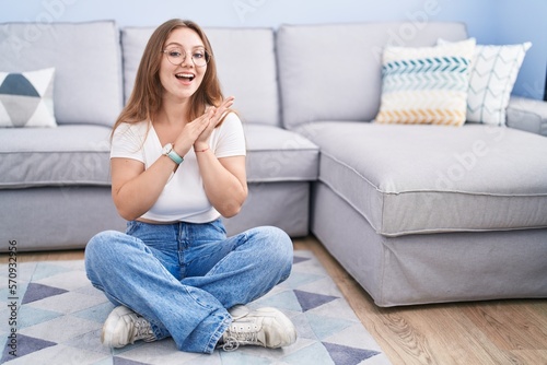 Young caucasian woman sitting on the floor at the living room clapping and applauding happy and joyful, smiling proud hands together