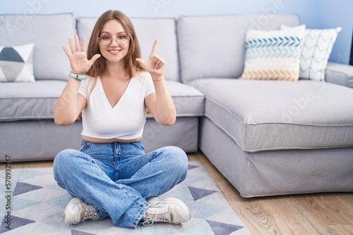 Young caucasian woman sitting on the floor at the living room showing and pointing up with fingers number seven while smiling confident and happy.