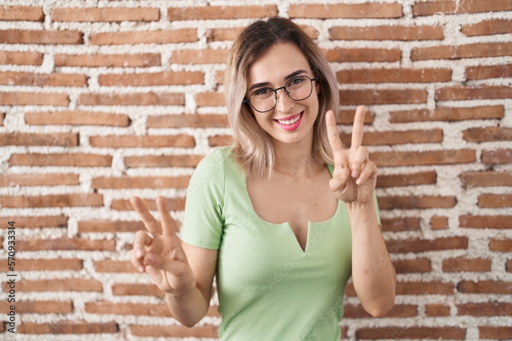 Young beautiful woman standing over bricks wall smiling looking to the camera showing fingers doing victory sign. number two. Young beautiful woman standing over bricks wall smiling looking to the camera showing fingers doing victory sign. number two.