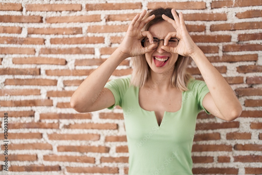 Young beautiful woman standing over bricks wall doing ok gesture like binoculars sticking tongue out, eyes looking through fingers. crazy expression. Young beautiful woman standing over bricks wall doing ok gesture like binoculars sticking tongue out, eyes looking through fingers. crazy expression.