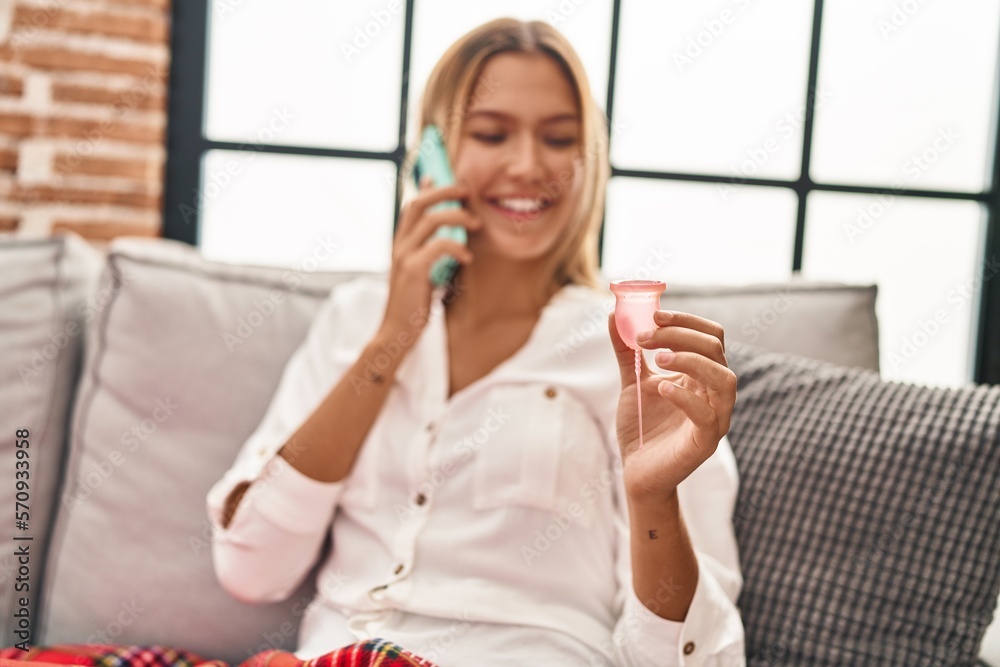 Young hispanic woman talking on smartphone holding menstrual cup at home Young hispanic woman talking on smartphone holding menstrual cup at home