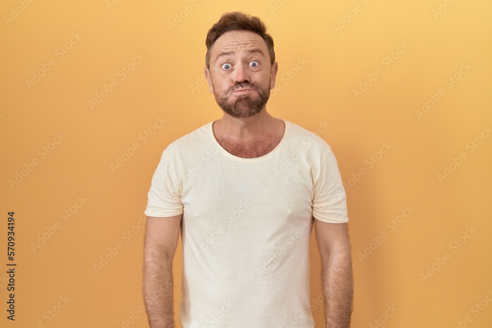 Middle age man with beard standing over yellow background puffing cheeks with funny face. mouth inflated with air, crazy expression. - obrazy, fototapety, plakaty 