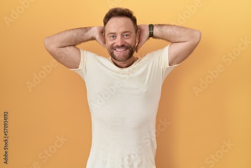 Middle age man with beard standing over yellow background relaxing and stretching, arms and hands behind head and neck smiling happy