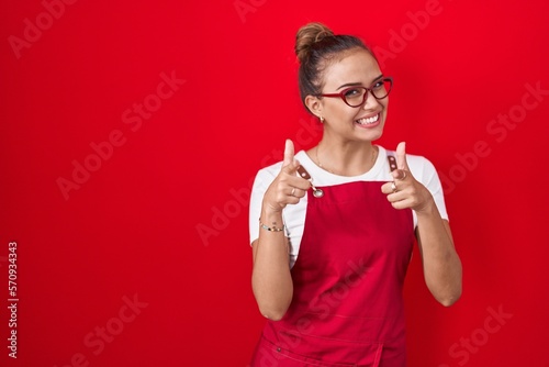 Fotografiet Young hispanic woman wearing waitress apron over red background pointing fingers to camera with happy and funny face