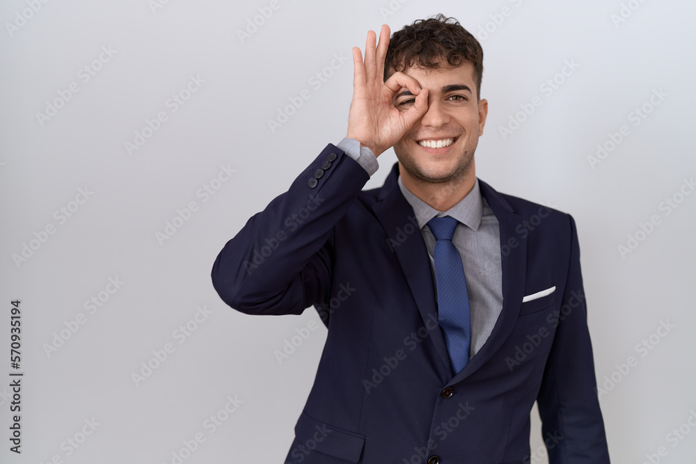 Young hispanic business man wearing suit and tie doing ok gesture with hand smiling, eye looking through fingers with happy face. Young hispanic business man wearing suit and tie doing ok gesture with hand smiling, eye looking through fingers with happy face.