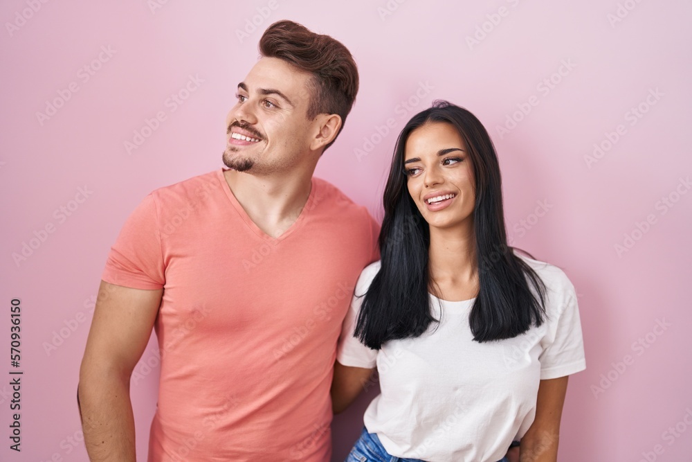 Young hispanic couple standing over pink background looking away to side with smile on face, natural expression. laughing confident. Young hispanic couple standing over pink background looking away to side with smile on face, natural expression. laughing confident.