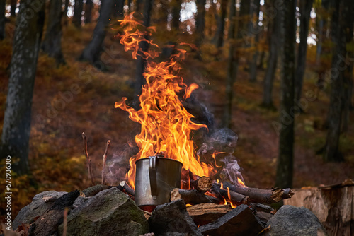 A metal mug is heated on an open fire. Strong fire on a blurred background of the autumn forest. Beautiful scene from tourist life