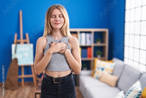 Blonde caucasian woman standing at living room smiling with hands on chest with closed eyes and grateful gesture on face. health concept.