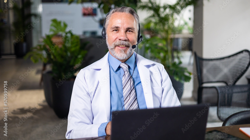 Looking at camera. Senior caucasian doctor online video call conference consult patient with headset on laptop computer. Doctor online consultation and telehealth medicine Looking at camera. Senior caucasian doctor online video call conference consult patient with headset on laptop computer. Doctor online consultation and telehealth medicine