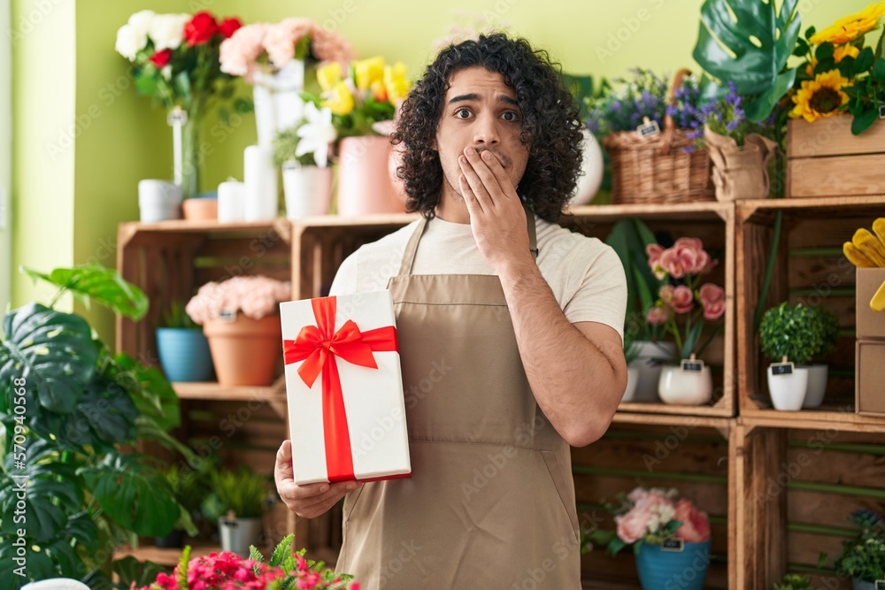 Hispanic man with curly hair working at florist shop holding gift covering mouth with hand, shocked and afraid for mistake. surprised expression