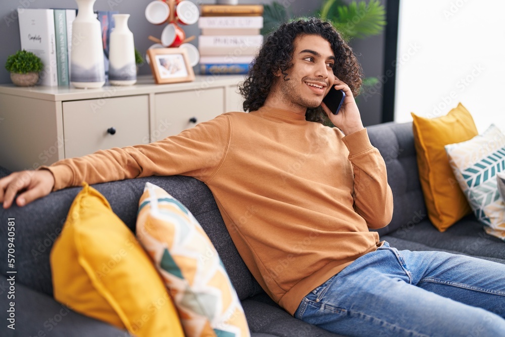 Young latin man talking on the smartphone sitting on sofa at home