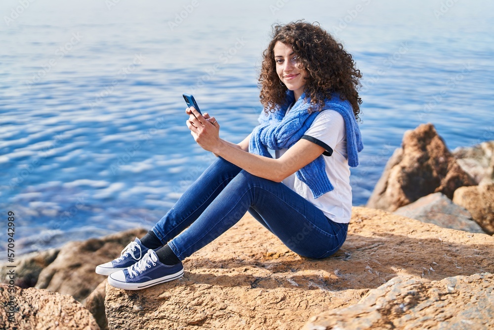 Young hispanic woman smiling confident using smartphone at seaside