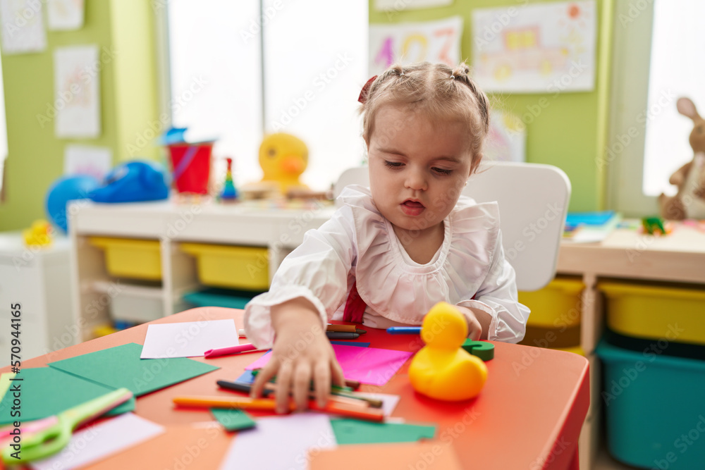 Adorable blonde toddler preschool student sitting on table drawing on paper at kindergarten Adorable blonde toddler preschool student sitting on table drawing on paper at kindergarten