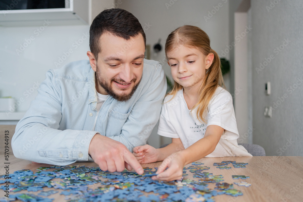 Fototapeta premium Father and daughter playing puzzles at home. Family connecting jigsaw puzzle pieces in a kitchen table. Kid assembling a jigsaw puzzle. Fun family leisure. Stay at home activity for kids.