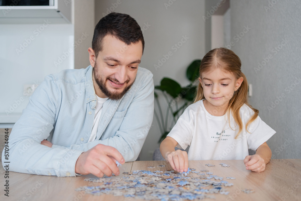 Fototapeta premium Father and daughter playing puzzles at home. Family connecting jigsaw puzzle pieces in a kitchen table. Kid assembling a jigsaw puzzle. Fun family leisure. Stay at home activity for kids.
