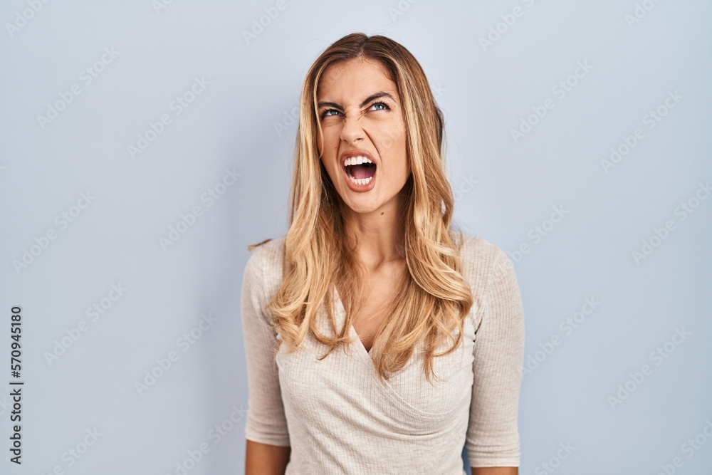 Young blonde woman standing over isolated background angry and mad screaming frustrated and furious, shouting with anger. rage and aggressive concept. Young blonde woman standing over isolated background angry and mad screaming frustrated and furious, shouting with anger. rage and aggressive concept.