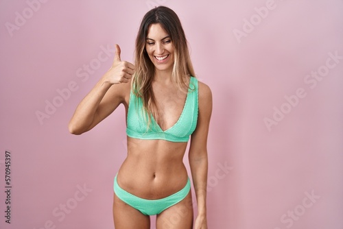 Young hispanic woman wearing bikini over pink background doing happy thumbs up gesture with hand. approving expression looking at the camera showing success.
