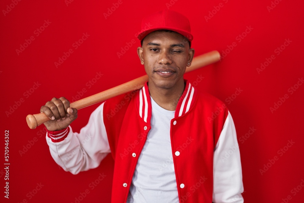Young hispanic man playing baseball holding bat winking looking at the camera with sexy expression, cheerful and happy face.