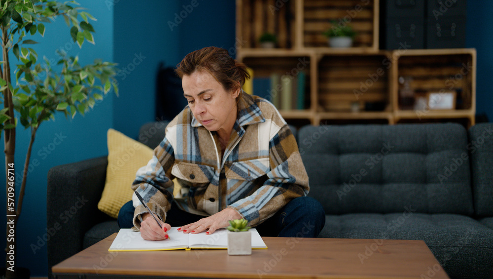 Middle age hispanic woman writing on notebook sitting on sofa at home Middle age hispanic woman writing on notebook sitting on sofa at home