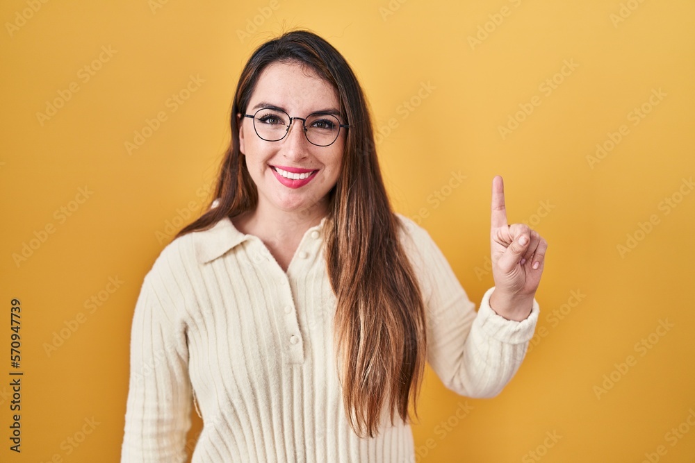 Young hispanic woman standing over yellow background showing and pointing up with finger number one while smiling confident and happy. Young hispanic woman standing over yellow background showing and pointing up with finger number one while smiling confident and happy.
