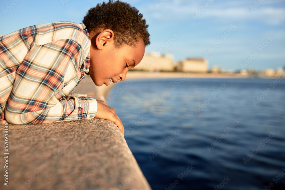 Side view of cute african american boy kid looking at blue water surface leaning on stoned fence of river embankment while walking down city streets in touristic place with copy pace on the right Side view of cute african american boy kid looking at blue water surface leaning on stoned fence of river embankment while walking down city streets in touristic place with copy pace on the right
