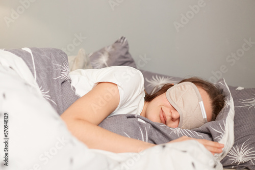 Indoor shot of young woman wearing sleepwear sleeping in a bed with eyes covered with gray mask, lying under blanket, can sleep longer during weekend. photo