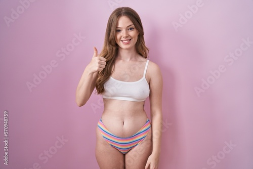 Caucasian woman wearing lingerie over pink background doing happy thumbs up gesture with hand. approving expression looking at the camera showing success. © Krakenimages.com