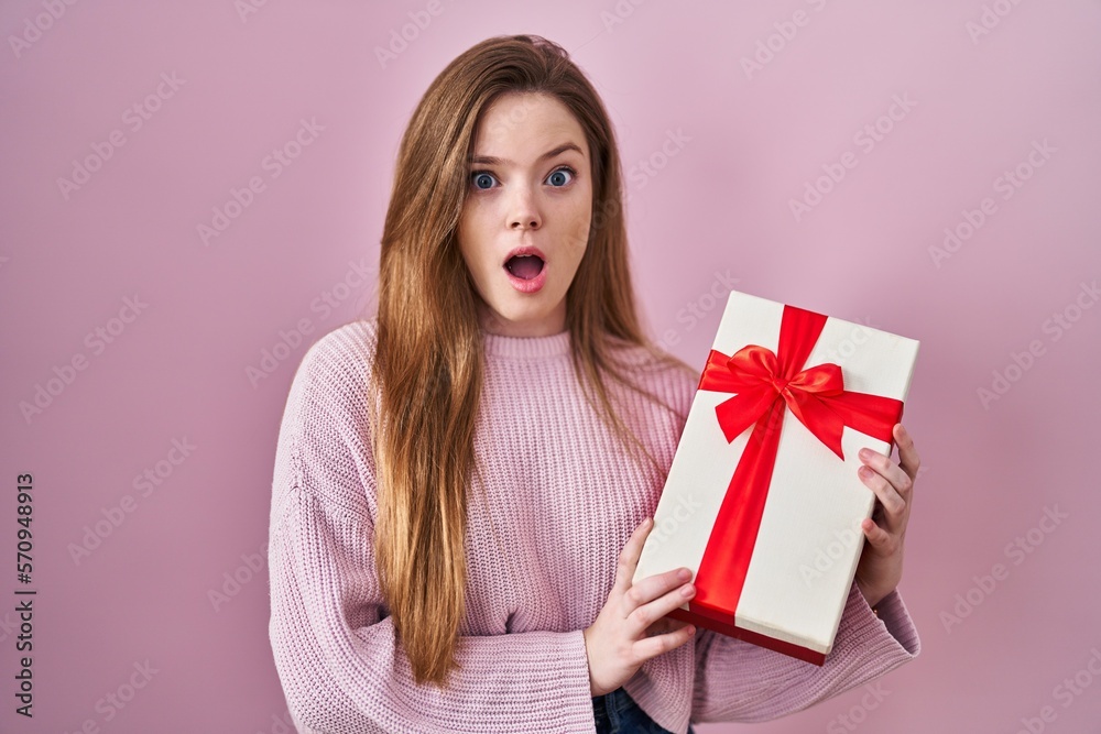 Young caucasian woman holding gift in shock face, looking skeptical and sarcastic, surprised with open mouth Young caucasian woman holding gift in shock face, looking skeptical and sarcastic, surprised with open mouth
