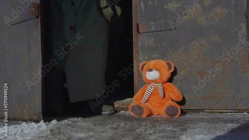 Soldier in antinuclear green suit and rubber gloves rise up abandoned children toy bear seated on frozen road surface outdoor. photo