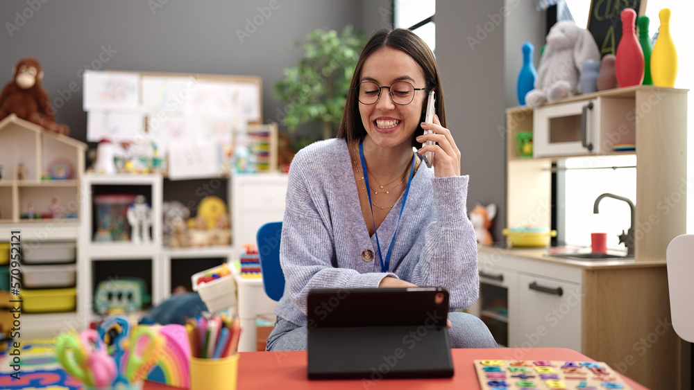 Young beautiful hispanic woman working as a teacher talking on smartphone using touchpad at kindergarten Young beautiful hispanic woman working as a teacher talking on smartphone using touchpad at kindergarten