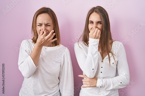 Middle age mother and young daughter standing over pink background smelling something stinky and disgusting, intolerable smell, holding breath with fingers on nose. bad smell