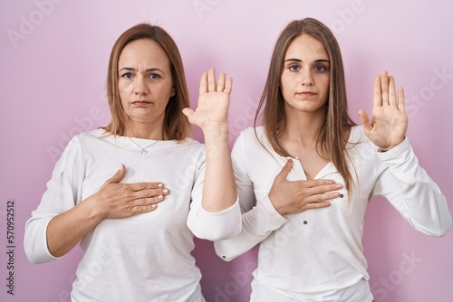 Middle age mother and young daughter standing over pink background swearing with hand on chest and open palm  making a loyalty promise oath