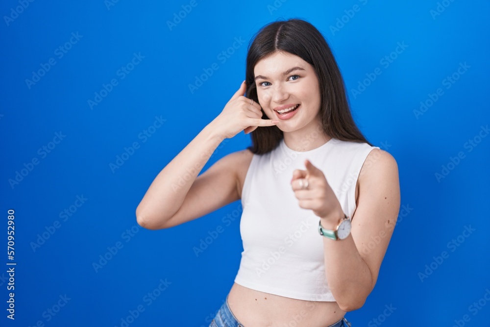 Young caucasian woman standing over blue background smiling doing talking on the telephone gesture and pointing to you. call me. Young caucasian woman standing over blue background smiling doing talking on the telephone gesture and pointing to you. call me.