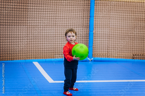 A small, beautiful boy, a child runs around the gym indoors, playing football with a green ball in his hands. Photography, sports. © shchus