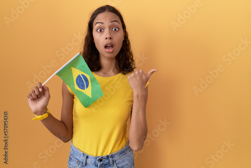 Young hispanic woman holding brazil flag surprised pointing with hand finger to the side  open mouth amazed expression.