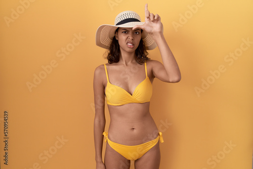 Young hispanic woman wearing bikini and summer hat making fun of people with fingers on forehead doing loser gesture mocking and insulting.