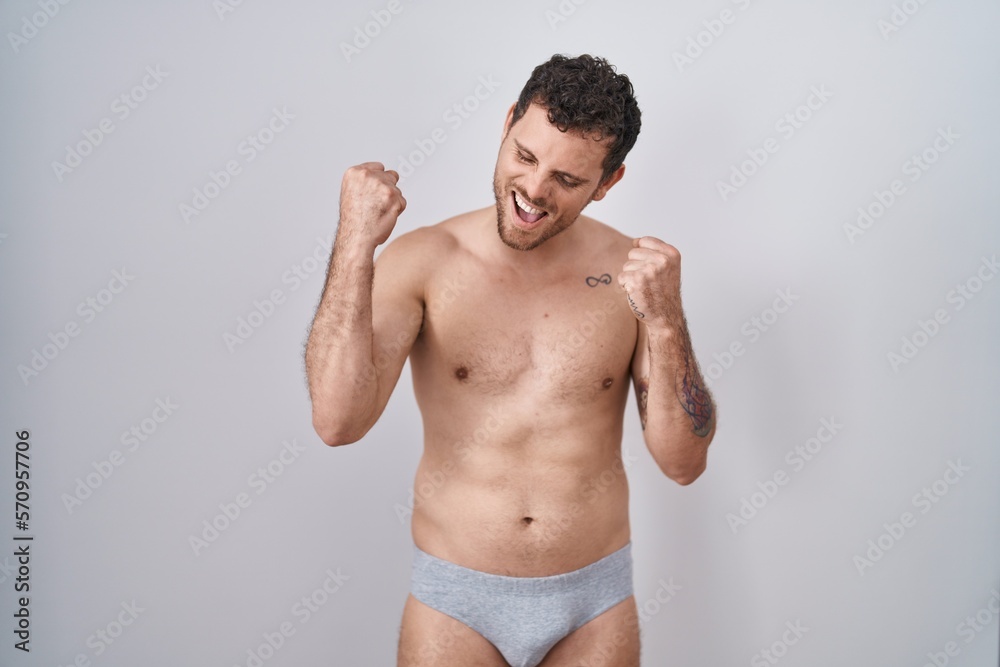 Young hispanic man standing shirtless wearing underware very happy and excited doing winner gesture with arms raised, smiling and screaming for success. celebration concept.