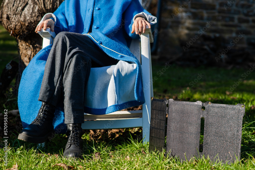 Fototapeta premium An unrecognizable woman sits in nature on green grass near a portable solar battery.