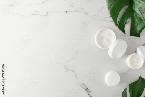 Skincare products concept. Flat lay photo of white cosmetic cream jars and green tropical leaves on marble background with empty space. Organic cosmetics idea.