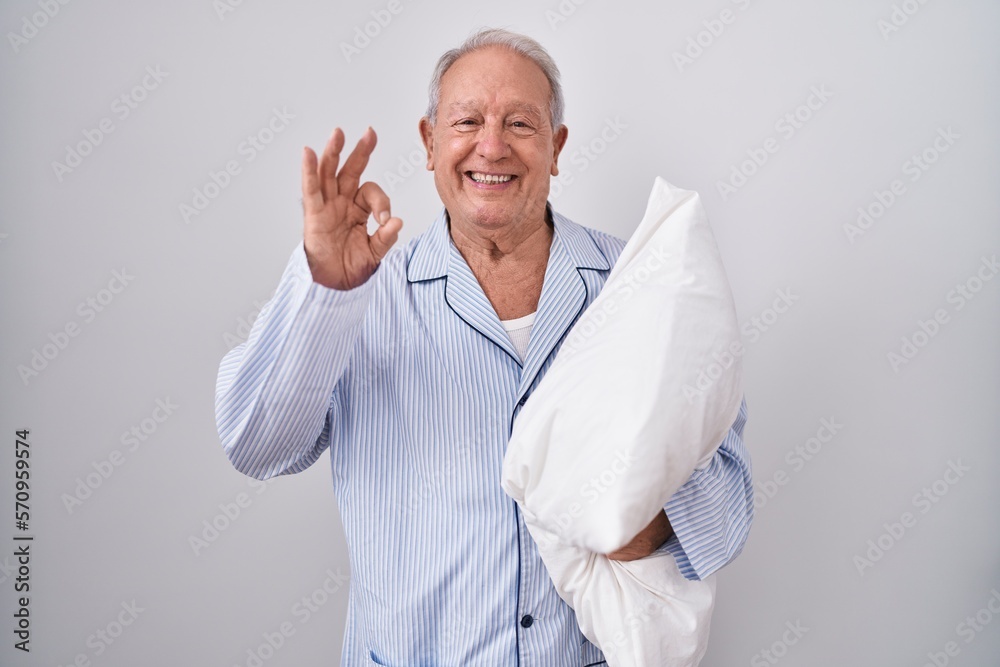Senior man with grey hair wearing pijama hugging pillow smiling positive doing ok sign with hand and fingers. successful expression. Senior man with grey hair wearing pijama hugging pillow smiling positive doing ok sign with hand and fingers. successful expression.