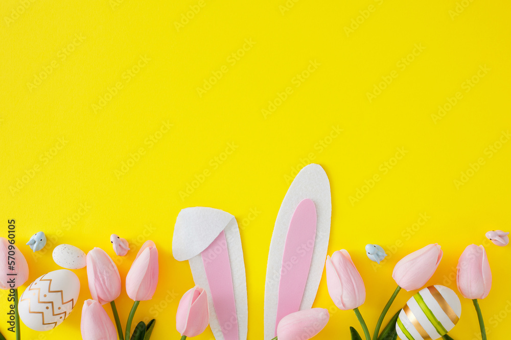 Easter decorations concept. Flat lay photo with bunny rabbit ears, tulips flowers and color eggs on yellow background with copyspace. Holiday invitation card idea.
