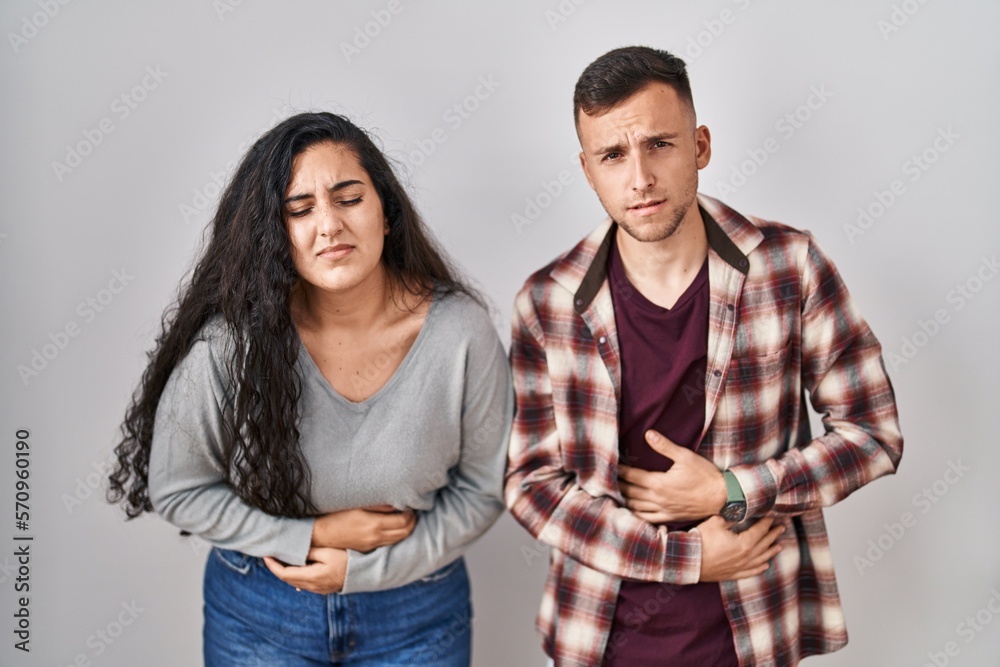 Young hispanic couple standing over white background with hand on stomach because indigestion, painful illness feeling unwell. ache concept. Young hispanic couple standing over white background with hand on stomach because indigestion, painful illness feeling unwell. ache concept.