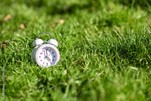 White color alarm clock on green grass. Place for text. Time, circadian rhythm, early rise concept.