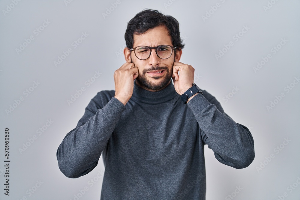 Handsome latin man standing over isolated background covering ears with fingers with annoyed expression for the noise of loud music. deaf concept. Handsome latin man standing over isolated background covering ears with fingers with annoyed expression for the noise of loud music. deaf concept.