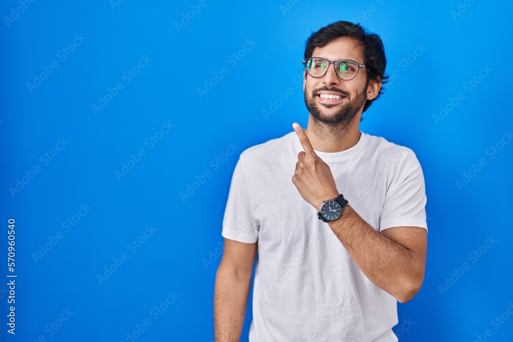 Handsome latin man standing over blue background cheerful with a smile of face pointing with hand and finger up to the side with happy and natural expression on face Handsome latin man standing over blue background cheerful with a smile of face pointing with hand and finger up to the side with happy and natural expression on face