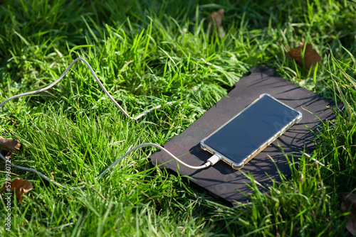 Portable solar battery and mobile phone on green grass. Charging electronic gadgets on a hike, during outdoor recreation. Ecological energy concept.
