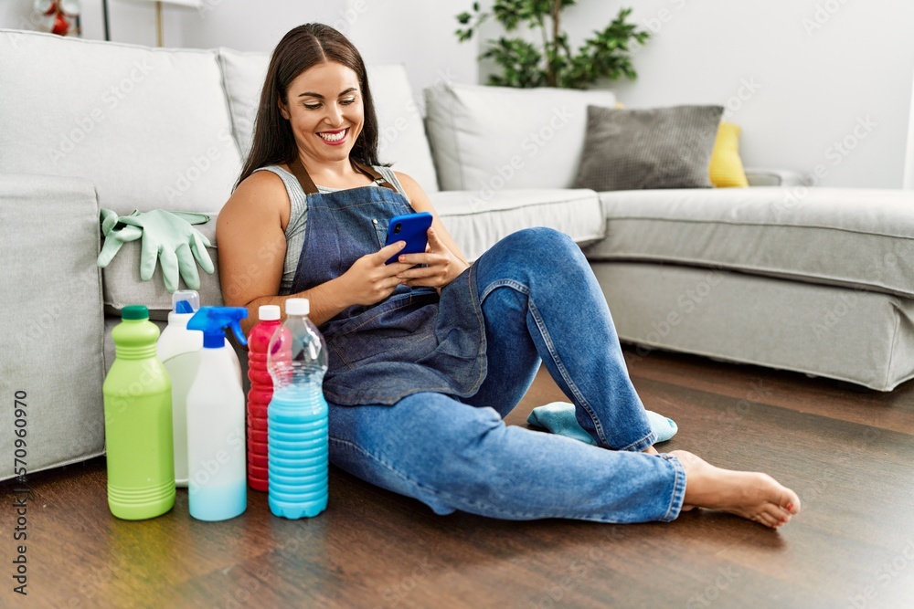 Young beautiful hispanic woman using smartphone sitting on floor with clean products at home Young beautiful hispanic woman using smartphone sitting on floor with clean products at home
